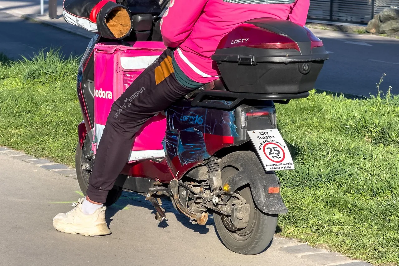Zusteller in pinker Jacke auf einem elektrischen Roller ohne Pedale, am Heck ein Schild »City Scooter – Ich bin ein Fahrrad – 25 km/h«.